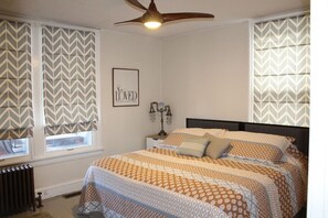 A bedroom with a bed, a ceiling fan, a framed picture on the wall, and patterned curtains.