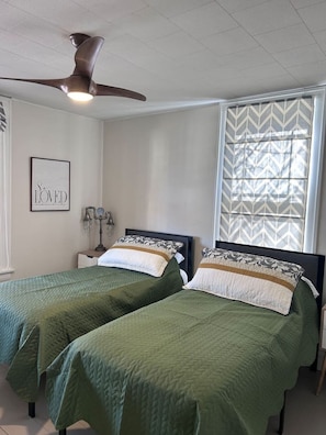 A bedroom with two single beds, a ceiling fan, a window with patterned blinds, and a framed 'LOVED' sign on the wall.