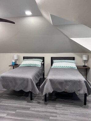 Une chambre moderne avec deux lits, chacun recouvert d’une couverture grise et agrémenté d’oreillers blancs. On y trouve des tables de chevet avec des lampes, et un petit objet bleu est posé sur l’une d’elles. La pièce est dotée d’un plafond en pente et d’un plancher en bois.