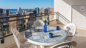 Uma varanda com mesa e cadeiras, um vaso com flores, e vista para a cidade e oceano.
