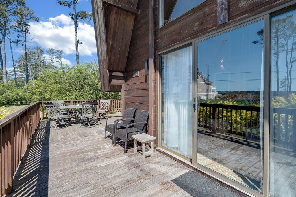 A wooden deck with outdoor furniture, a glass door leading to a balcony, and a view of trees and sky.