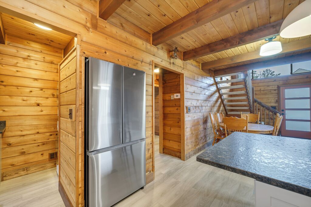 A wooden cabin interior with a kitchen area featuring a stainless steel refrigerator, a dining table with chairs, and a staircase leading to an upper level.