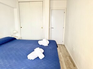 Quarto de hotel com uma cama grande, duas toalhas brancas, uma mesinha de cabeceira e uma porta branca.