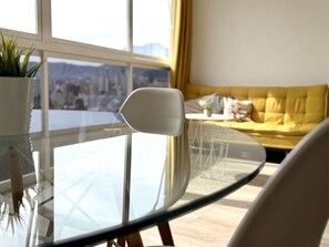 Uma sala de estar moderna com uma mesa de vidro, uma cadeira branca, um sofá amarelo e vista para o horizonte urbano através de grandes janelas.