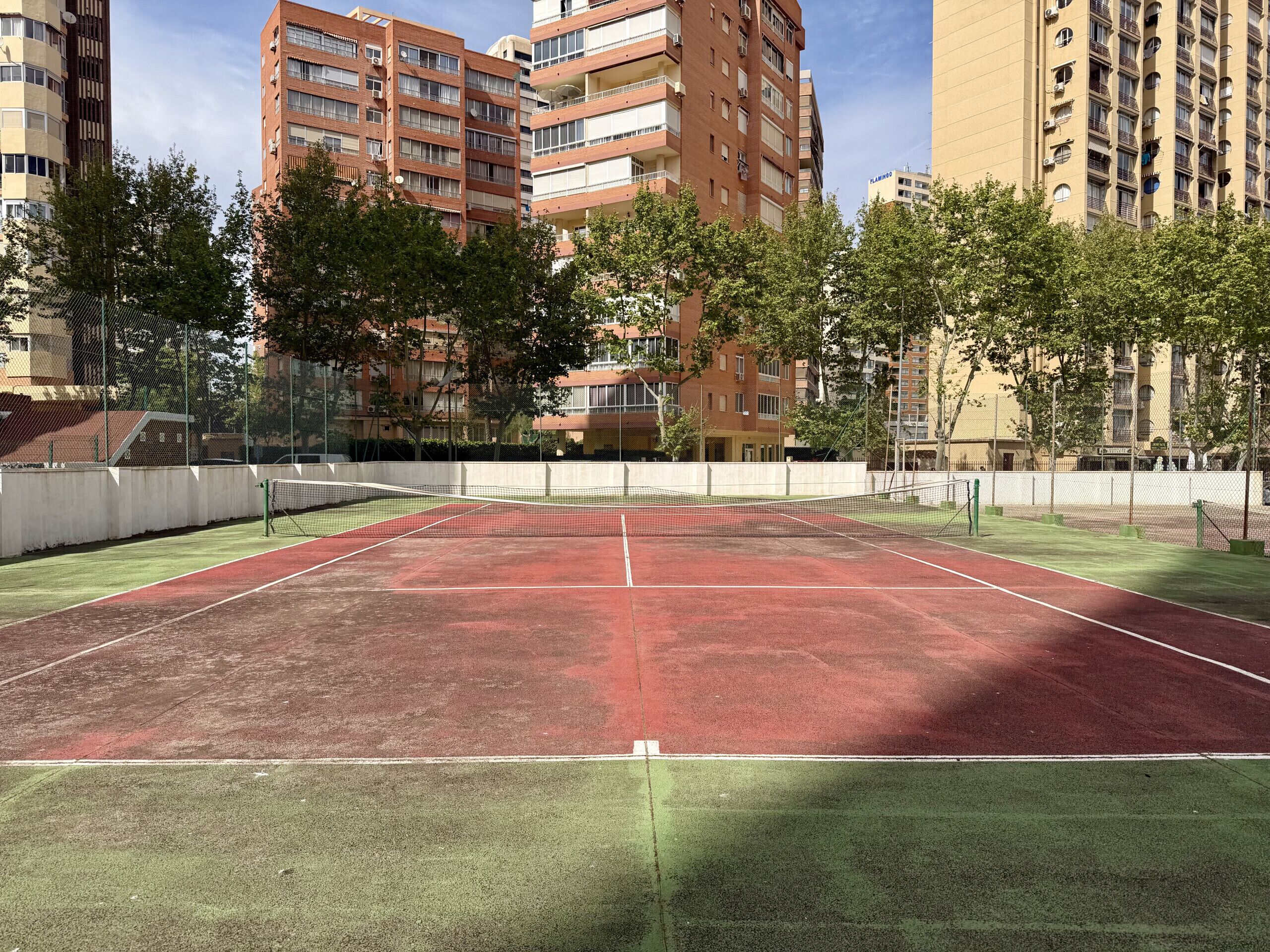 En tennisbana omgiven av träd och höga byggnader.