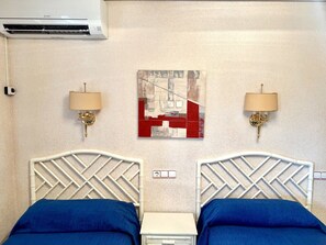 Quarto de hotel com duas camas, ar condicionado fixado na parede, duas arandelas, uma mesinha de cabeceira e uma obra de arte abstrata na parede.