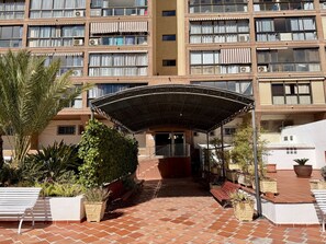 Un edificio de varios pisos con un acceso cubierto, asientos al aire libre y plantas en macetas.
