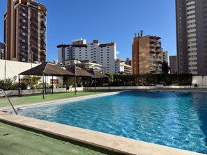 Piscina com superfície de água azul cristalina, rodeada por um gramado verde e área sombreada com telhado de palha.