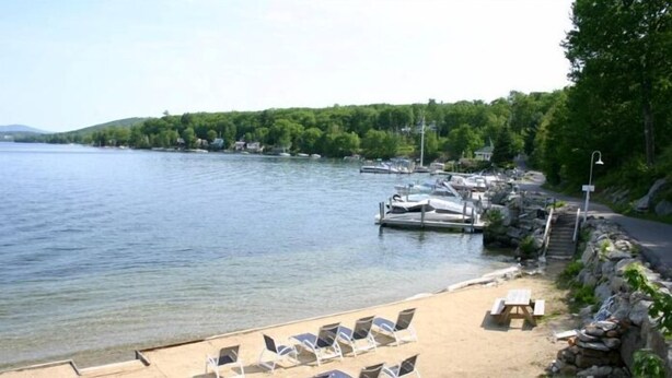 Vista a un lago con barcos amarrados, una playa de arena con sillas de descanso y un muro de piedra.