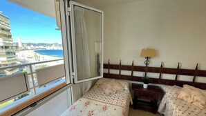 Hotellihuone, jossa on parveke, kukallinen vuodevaatteisto, yöpöytä lampun kanssa ja näkymä rannalle ja kaupunkiin.