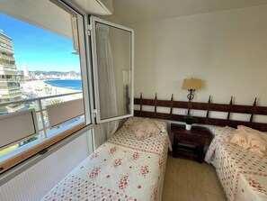A hotel room with a balcony, a bed with floral bedding, a bedside table with a lamp, and a view of the beach and cityscape. - Seafront dream apartment (Benidorm)