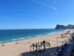 A beach with a clear view of the ocean, a road with parked cars, and a distant hill with buildings. - Seafront dream apartment (Benidorm)