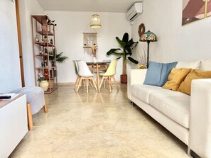 Uma sala de estar com um sofá branco, uma mesa de jantar com cadeiras, uma estante de livros e um vaso com planta.