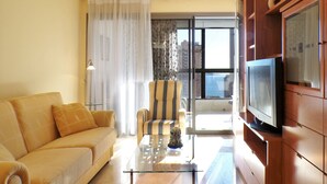 Una sala de estar con sofá, sillón, mesa de centro de vidrio y televisor sobre un mueble de madera.