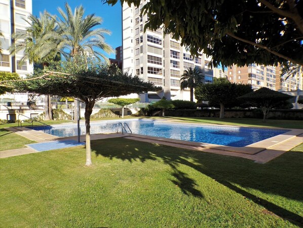 A swimming pool surrounded by well-maintained grass and trees, with modern buildings in the background.