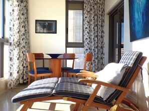 A room with a wooden dining table, blue cushioned chairs, a wooden lounge chair with a striped cushion, and floral curtains.