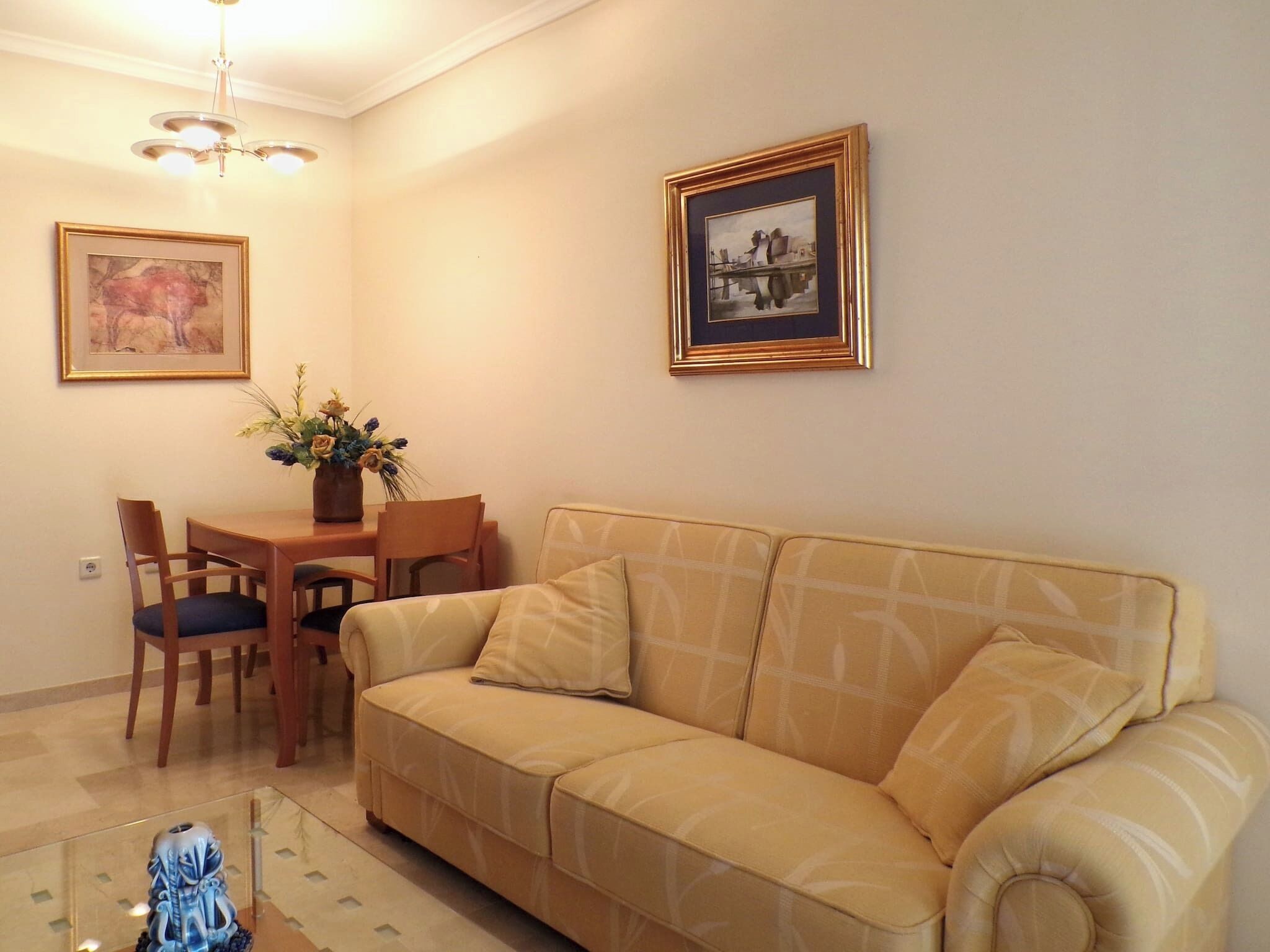 A living room with a beige sofa, a dining table with chairs, a vase of flowers, and two framed pictures on the wall.