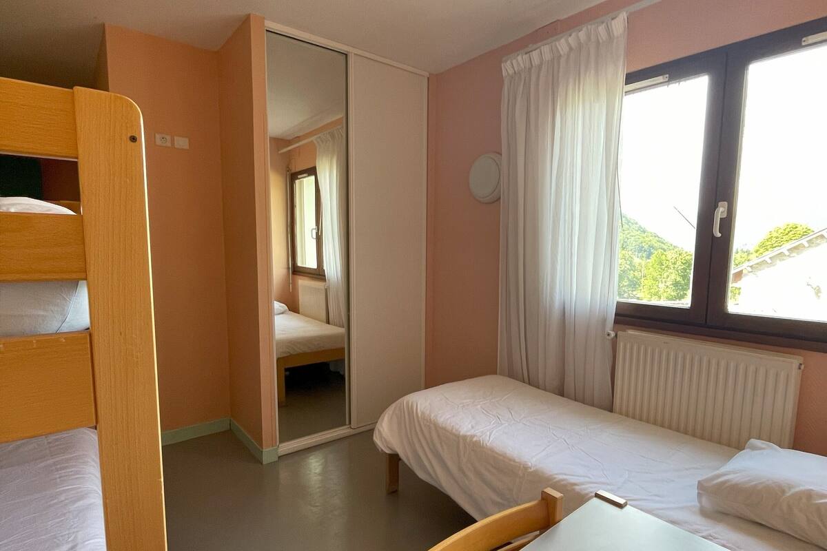 A dormitory room with bunk beds, a single bed, a table, and a window with curtains.