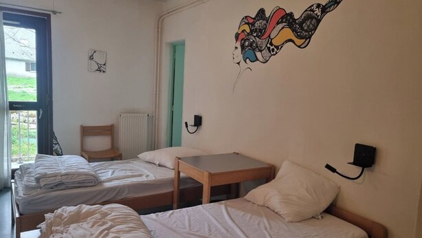 A dormitory room with two single beds, a small wooden table, a wall-mounted lamp, and a window with a view of greenery outside. - La Baleine Blanche (Campan)