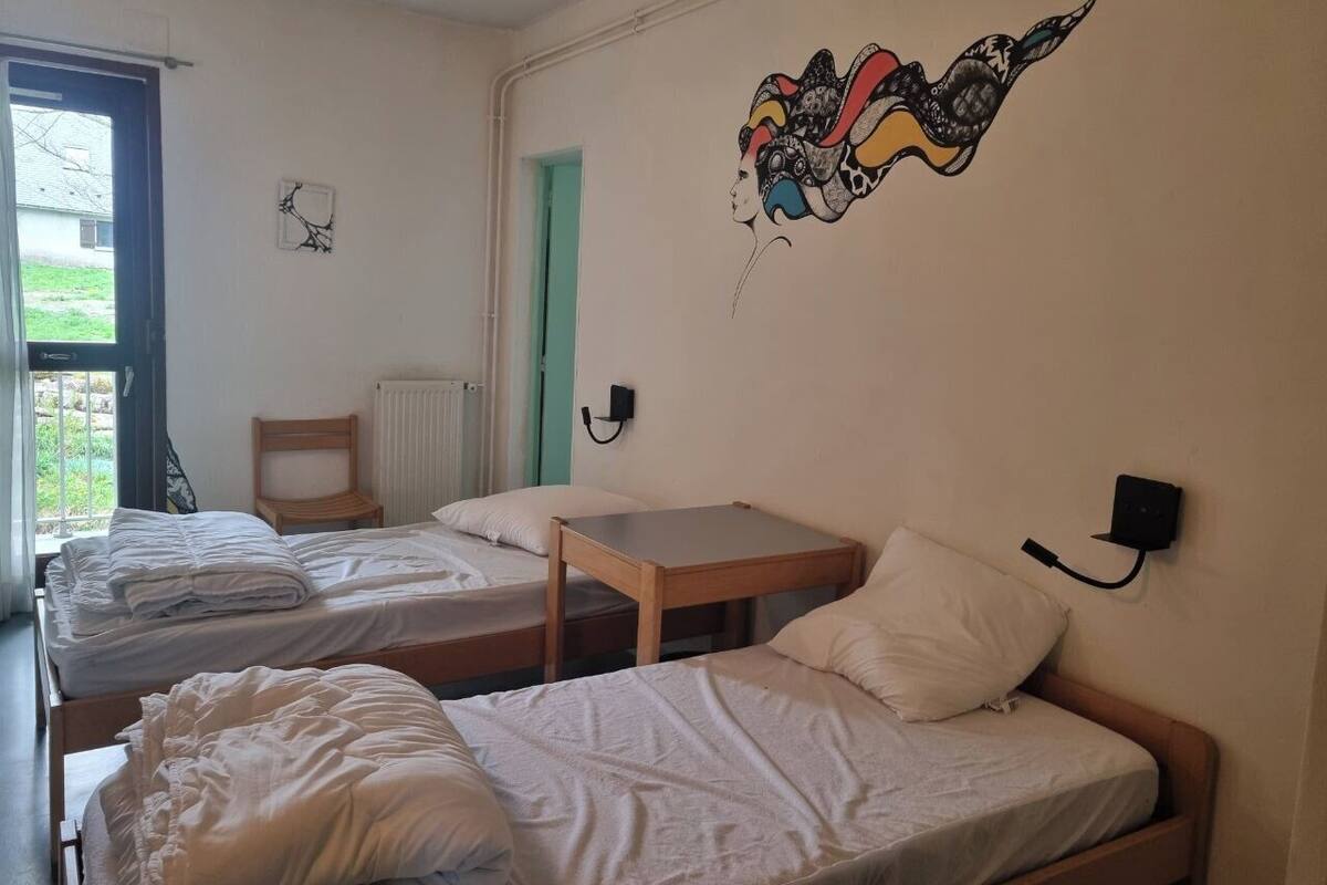 A dormitory room with two single beds, a small wooden table, a wall-mounted lamp, and a window with a view of greenery outside.