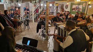 A room with a piano, a sewing machine, and a group of people gathered around tables, possibly in a communal area. - La Baleine Blanche (Campan)