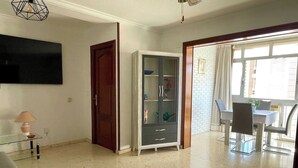 Een ruime woonkamer met een flatscreen televisie aan de muur, een glazen salontafel met een lamp, een houten deur en een eethoek met een tafel en stoelen.