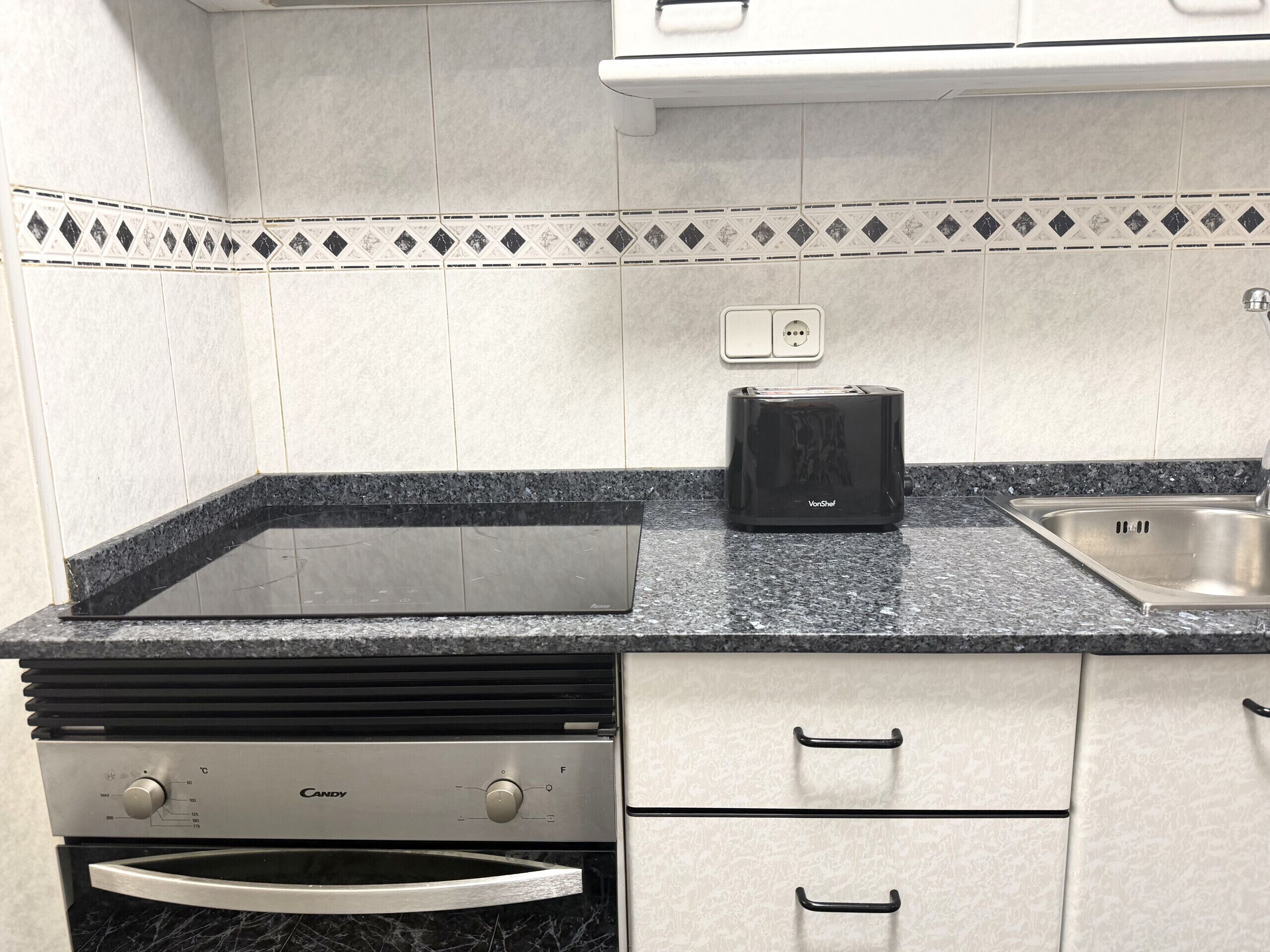 Een keuken met een zwarte broodrooster, een roestvrijstalen spoelbak, een vaatwasser en een betegelde spatwand met een patroon.