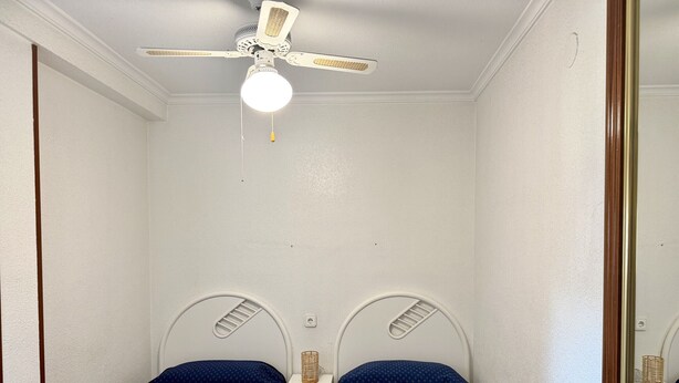 Une petite chambre propre, avec deux lits simples, un ventilateur de plafond et un miroir.