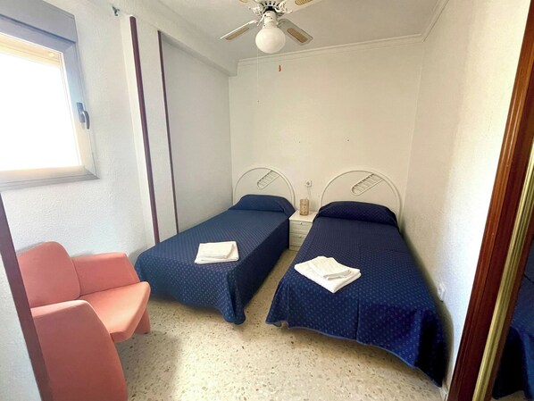 Une petite pièce avec deux lits simples, une chaise, un ventilateur de plafond et une fenêtre.