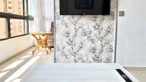 Une chambre moderne avec un téléviseur à écran plat fixé sur un mur de marbre, une petite table ronde avec une chaise et une télécommande posée sur une surface blanche.