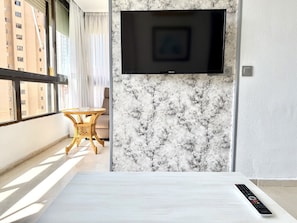 Een moderne kamer met een flatscreen televisie aan een marmeren wand, een klein rond tafeltje met een stoel, en een afstandsbediening op een witte ondergrond.