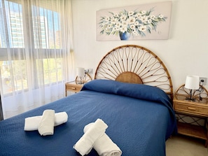 Een net opgemaakt bed met blauw beddengoed, een houten hoofdbord, een nachtkastje met een lamp en een schilderij van bloemen aan de muur.
