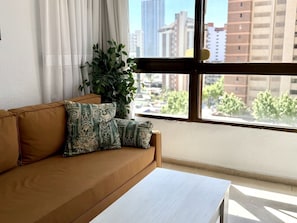 Een kamer met een bank, een tafel, een plant en een raam met uitzicht over de stad.
