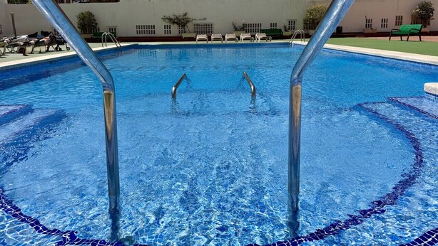 Une piscine avec des échelles en métal, entourée d’un mur blanc et de quelques chaises longues.