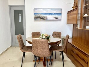 Een eetgedeelte met een ronde tafel, vier stoelen, een vaas met bloemen en een schilderij van een strandgezicht.
