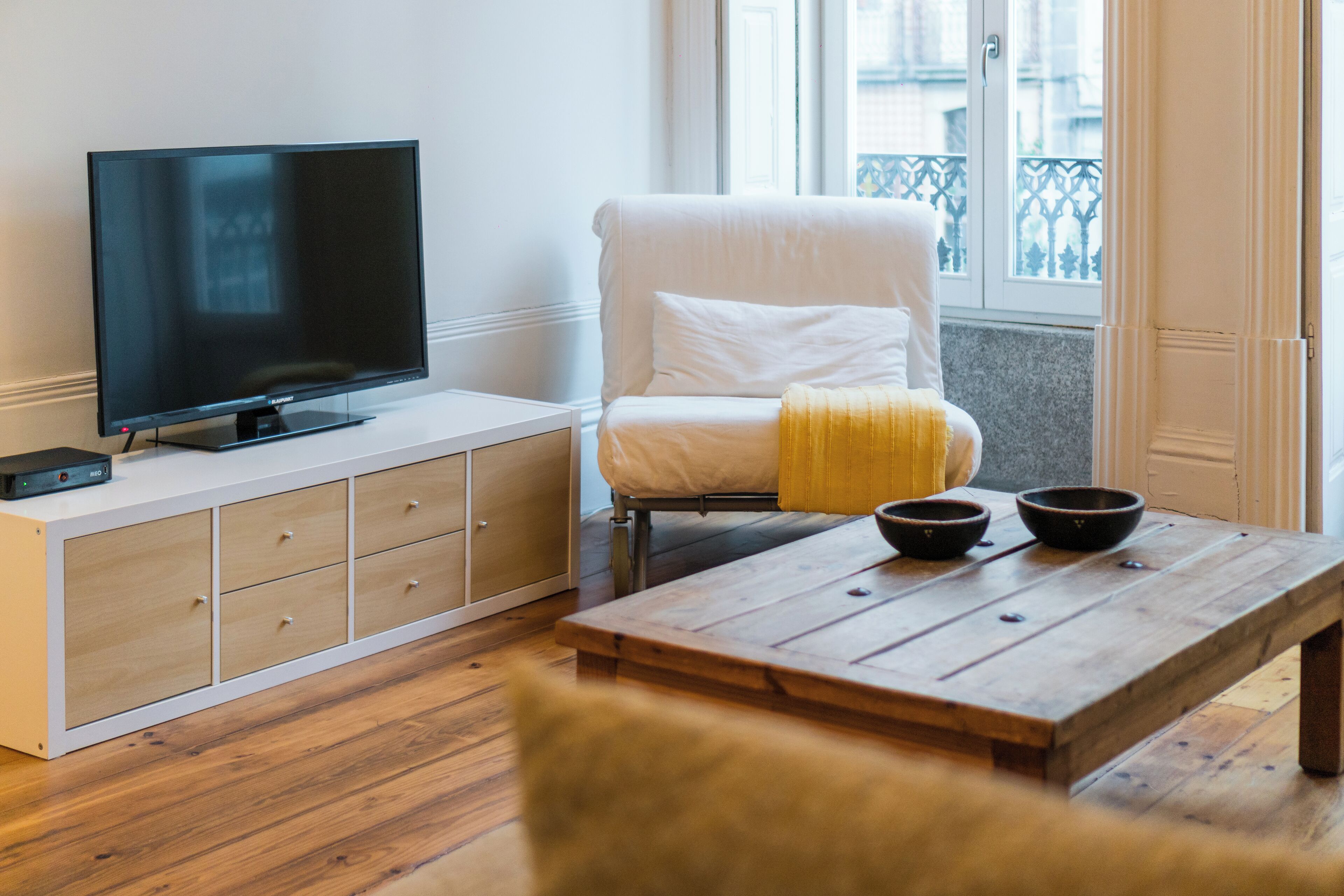 A living room with a flat-screen TV on a white cabinet, a white armchair with a yellow throw, and a wooden coffee table with two bowls.