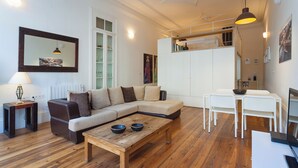 Ein modernes Wohnzimmer mit Ecksofa, einem Holztisch, einem Esstisch mit Stühlen und einer darüberliegenden Küchenzeile.