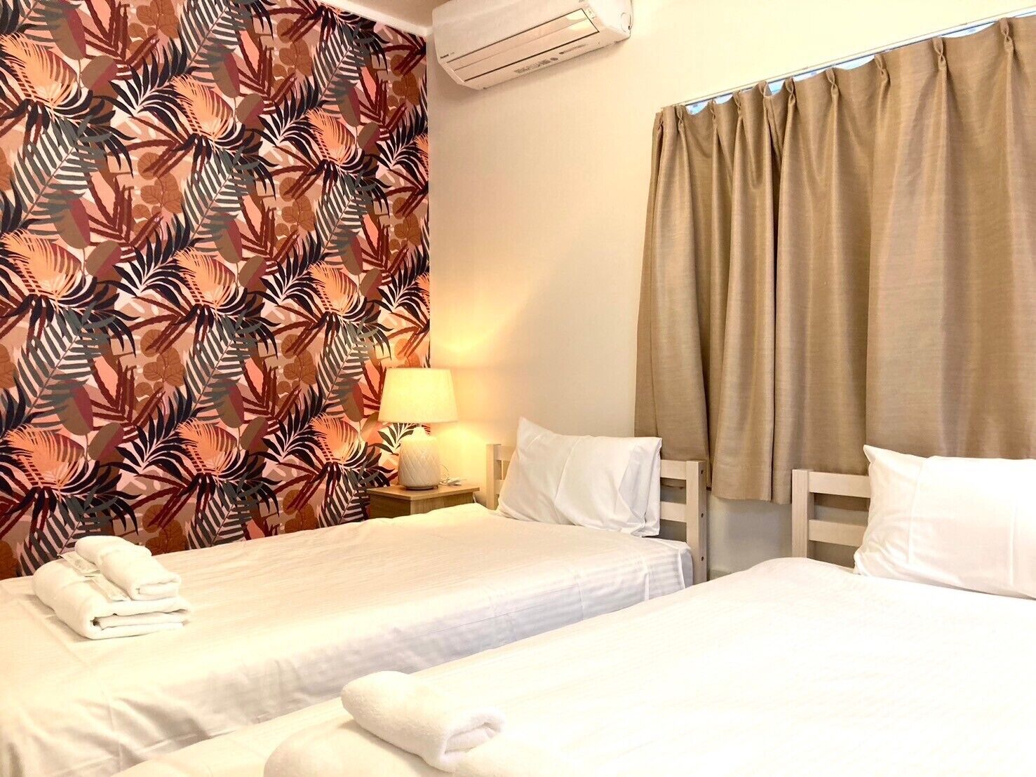 Habitación de hotel con dos camas, una mesita de noche con lámpara y cortina estampada con motivos tropicales.