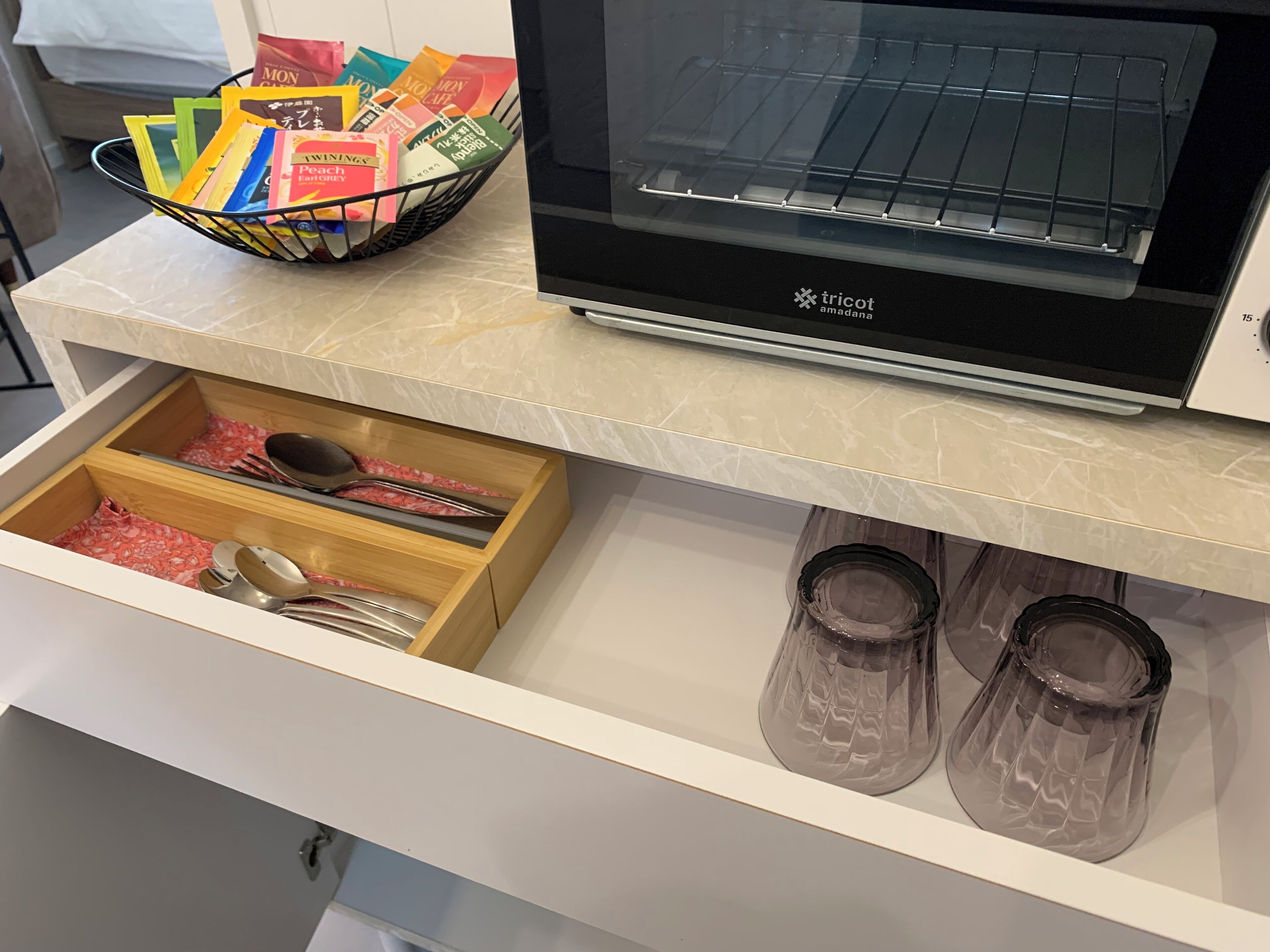 Una cocina con microondas, horno tostador, un cajón con utensilios de cocina y una cesta con productos envasados y de colores.