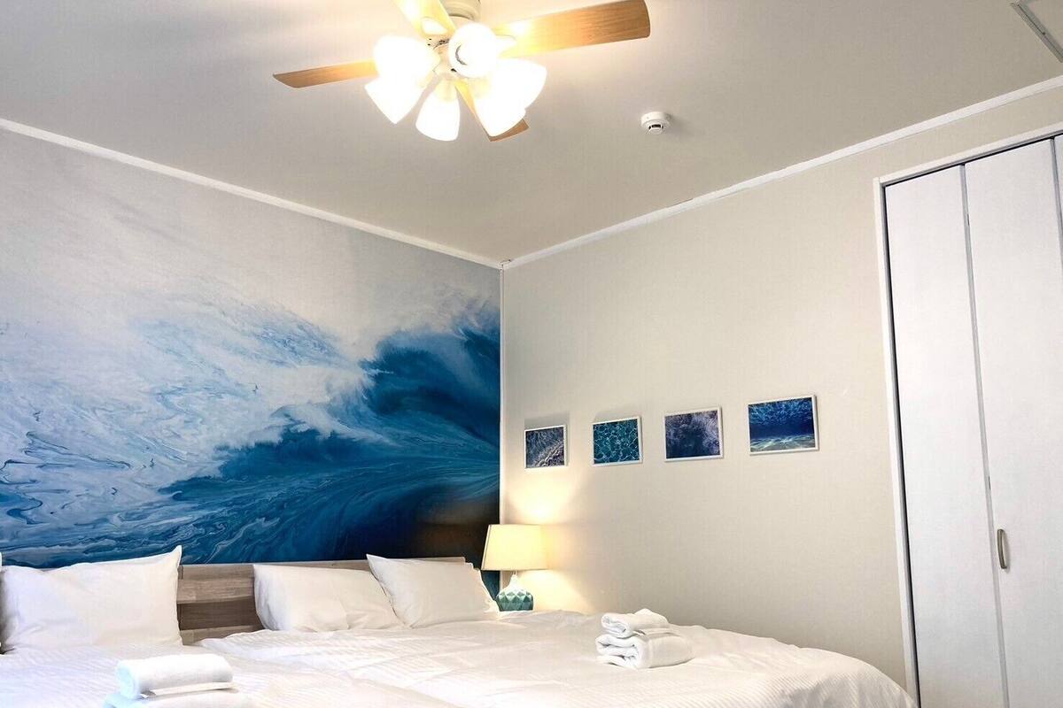 Una habitació d'hotel amb un llit, un ventilador de sostre, una paret amb un gran quadre d'una onada marina i quatre petites fotografies emmarcades a la paret.