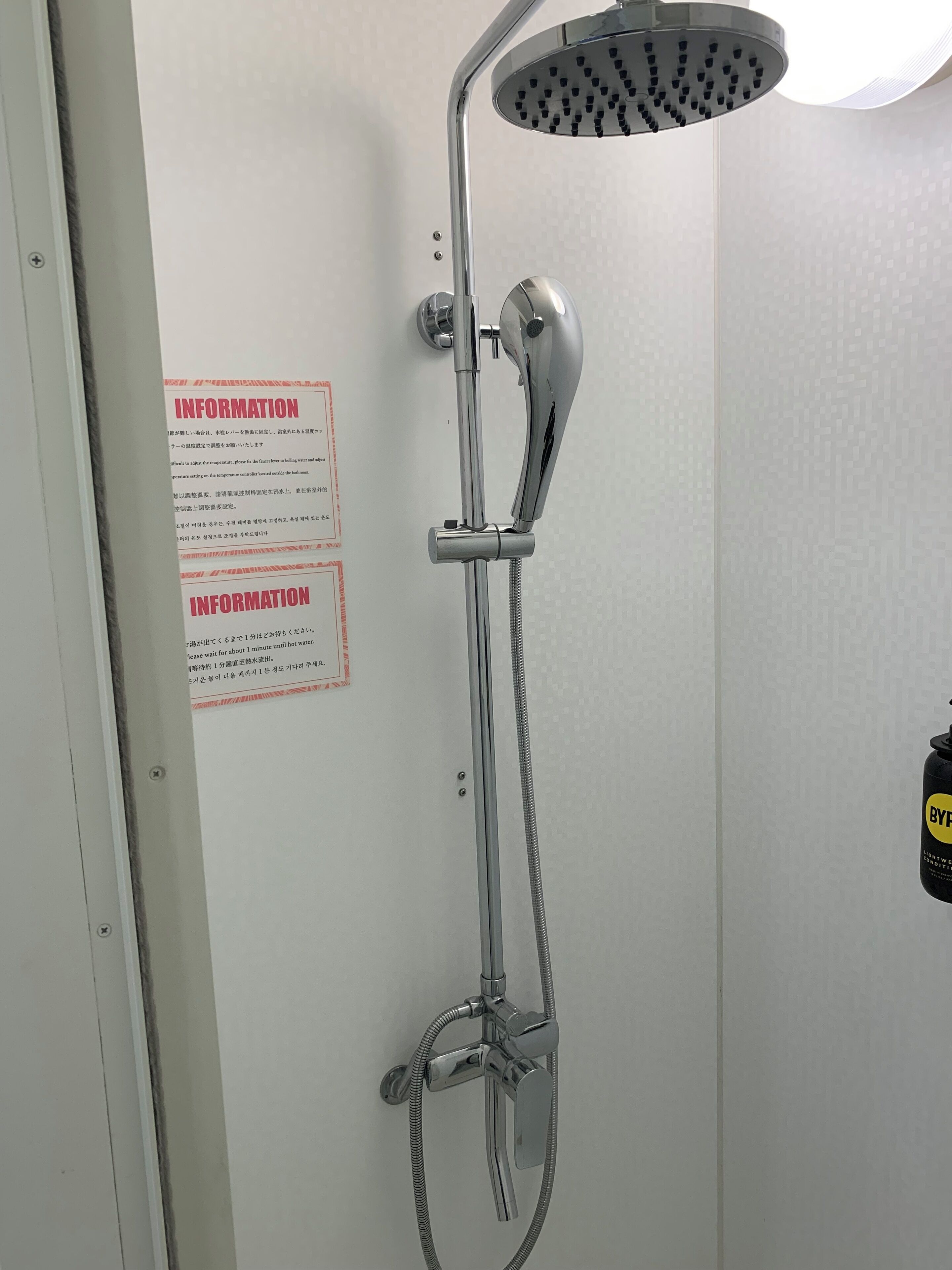 A shower with a handheld showerhead and a wall-mounted control. There is a sign with information on the wall.