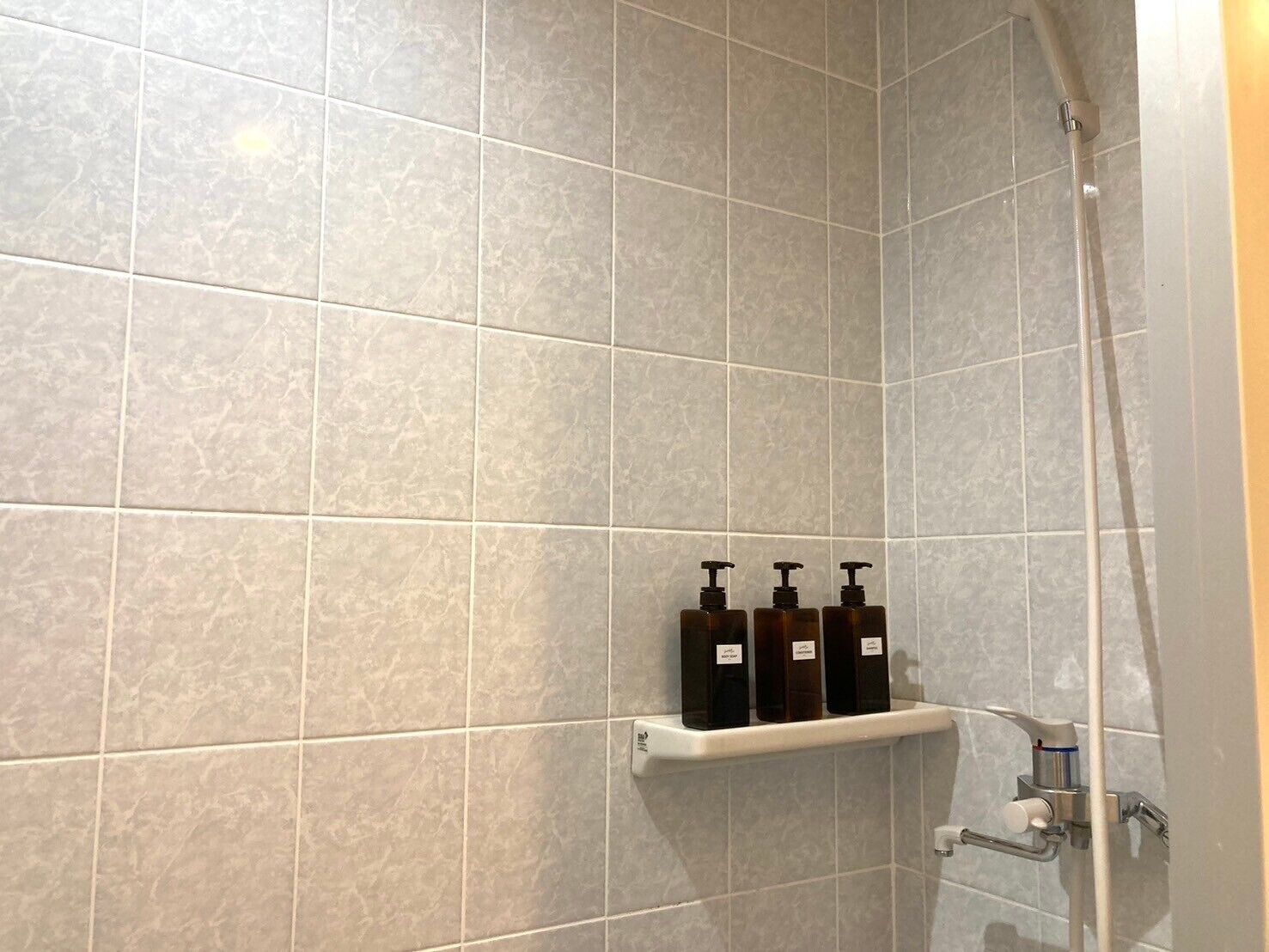 A shower area with grey tiles, a shelf with three bottles, and a handheld showerhead.