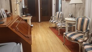 Een hotelkamer met een houten bureau, een klein rond tafeltje, twee gestreepte fauteuils, een rood tapijt en een schilderij aan de muur.