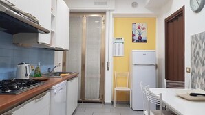 A compact kitchen with white cabinets, a gas stove, a dishwasher, a refrigerator, and a dining area with a white table and chairs. - Le Campane Apartment Verona Center (VERONA)