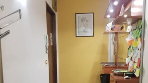 A hallway with a yellow wall, a framed picture, a trash bin, and a desk with papers and a basket. - Le Campane Apartment Verona Center (VERONA)