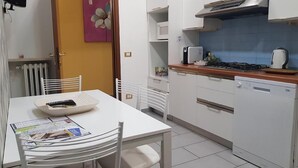 A compact kitchen with white cabinets, a stove, and a microwave. There is a dining table with chairs and a bowl. A flower painting is on the wall. - Le Campane Apartment Verona Center (VERONA)