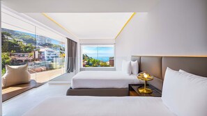 A modern hotel room with a large balcony, two beds, a bedside table with a lamp, and a view of the city and sea.