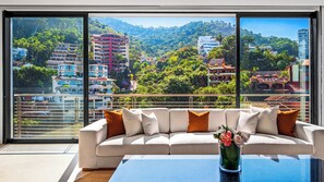 A modern living room with a large window offering a view of a hillside with buildings and greenery.
