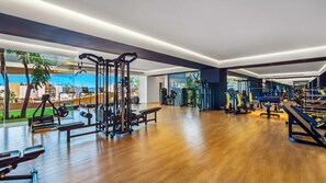 A spacious gym with various exercise equipment, large windows offering views of the outdoors, and a wooden floor.