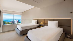Una habitación de hotel moderna con dos camas, una mesita de noche con lámpara y un ventanal con vistas a la ciudad.
